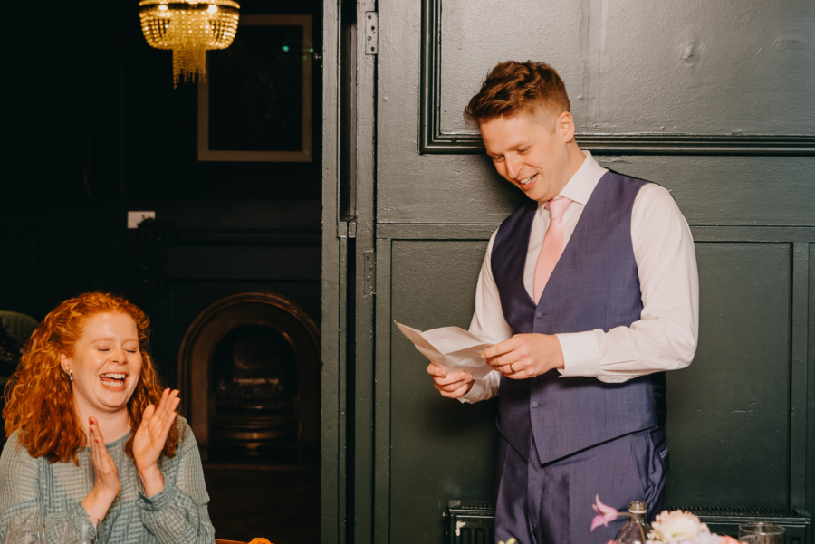 Groom giving a speech while a guest laughs, captured through candid wedding photography.